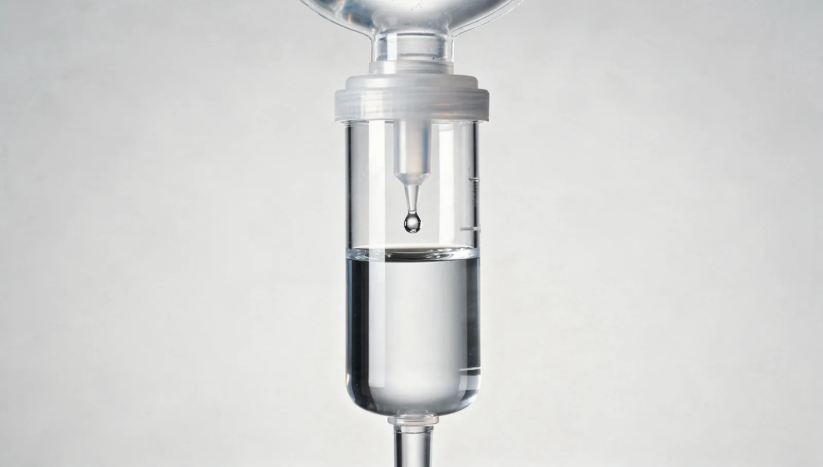 A clear rigid cylindrical IV drip chamber hanging below the spike of a fluid bag, filled a third of the way with clear fluid, a single drop hanging from the small inner spike in the air gap above the fluid line, close against a plain pale backdrop