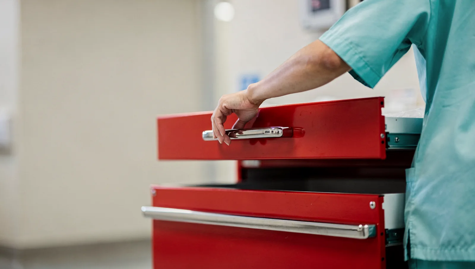 A person in surgical scrubs and a cap pulling open the drawer of a red emergency cart at speed, close on the drawer and one hand