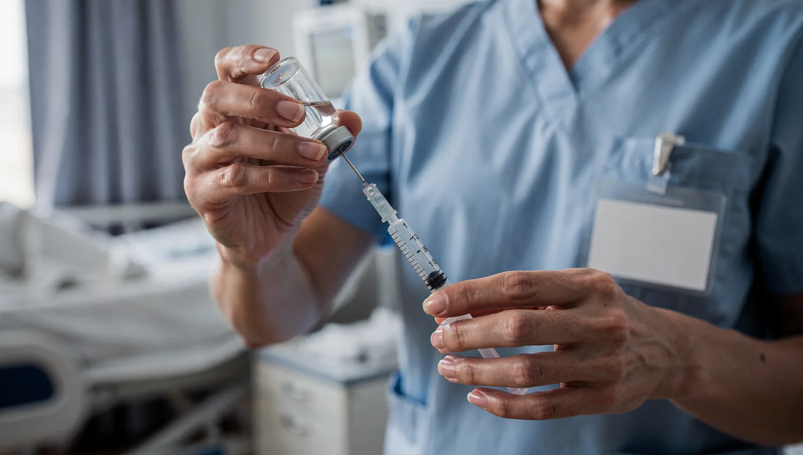 A close view of a nurse's hands drawing medication up into a syringe from a small glass vial, the needle inserted through the vial's rubber stopper.